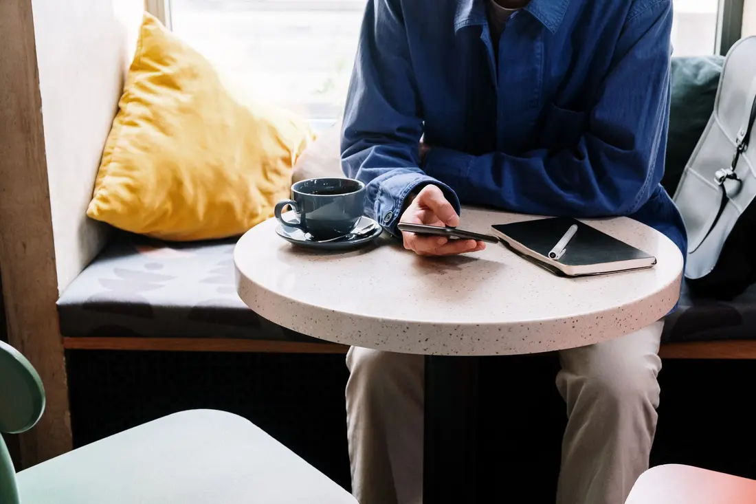A person sitting at a cafe table typing on a phone next to a cup of coffee