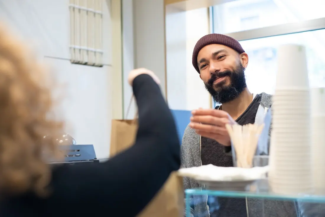A shop owner behind the counter handing a paper bag to a customer