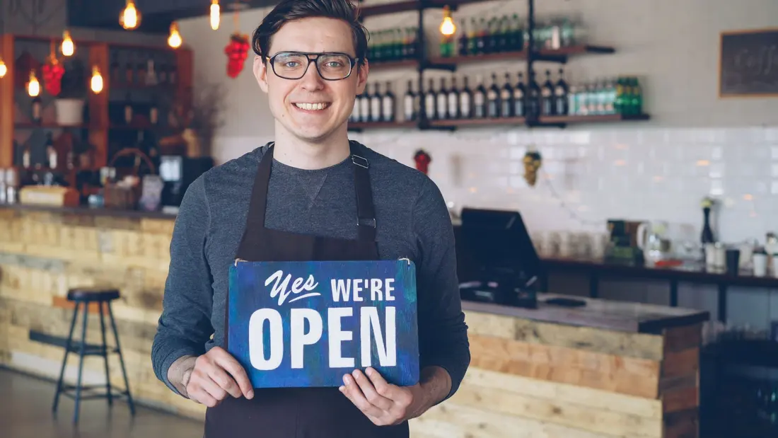 A shop owner standing in his doorway holding a Yes We Are Open sign